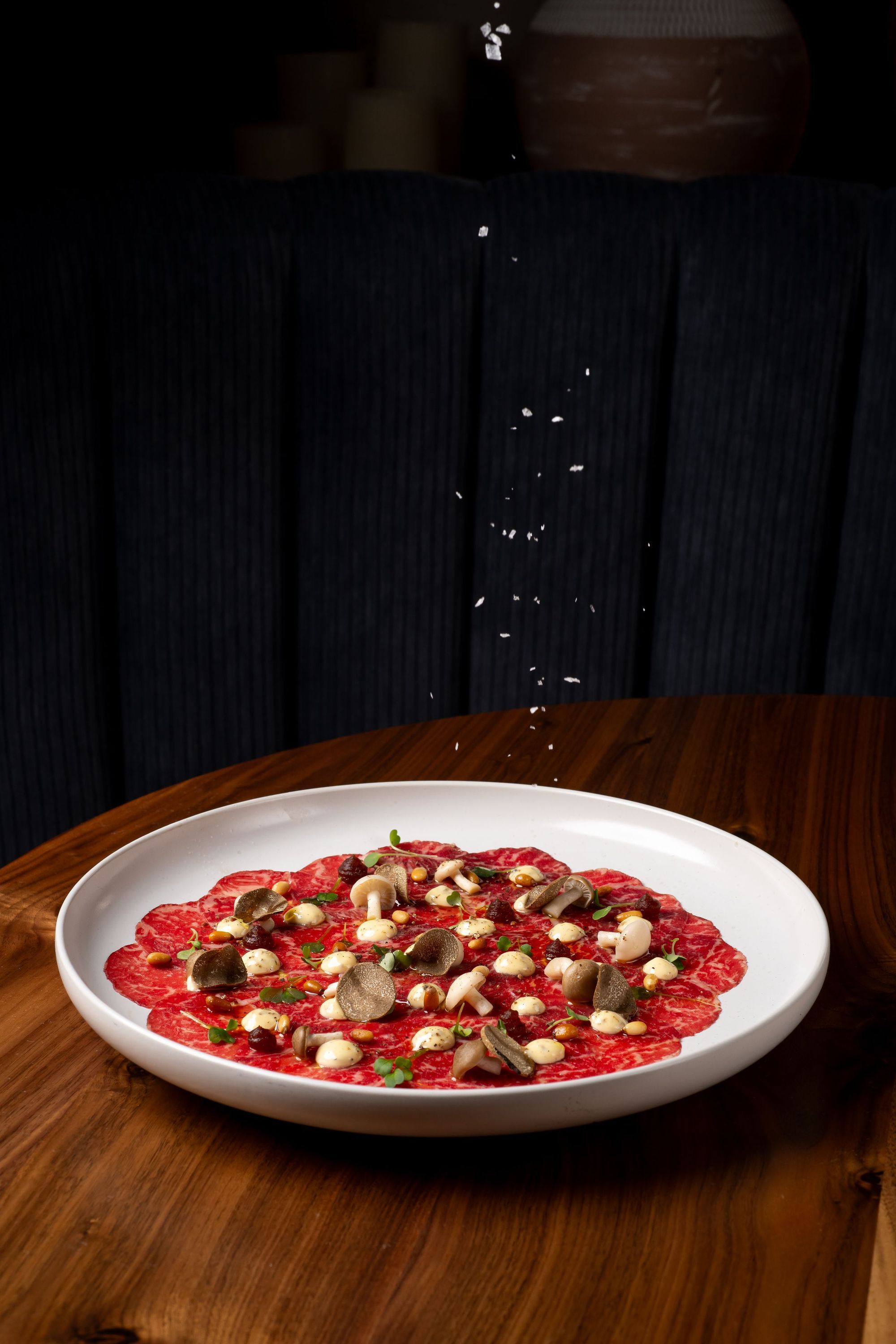 Thinly sliced beef carpaccio on a white plate with mushrooms and creamy sauce, on a wooden table at Sparrow Italia Miami.