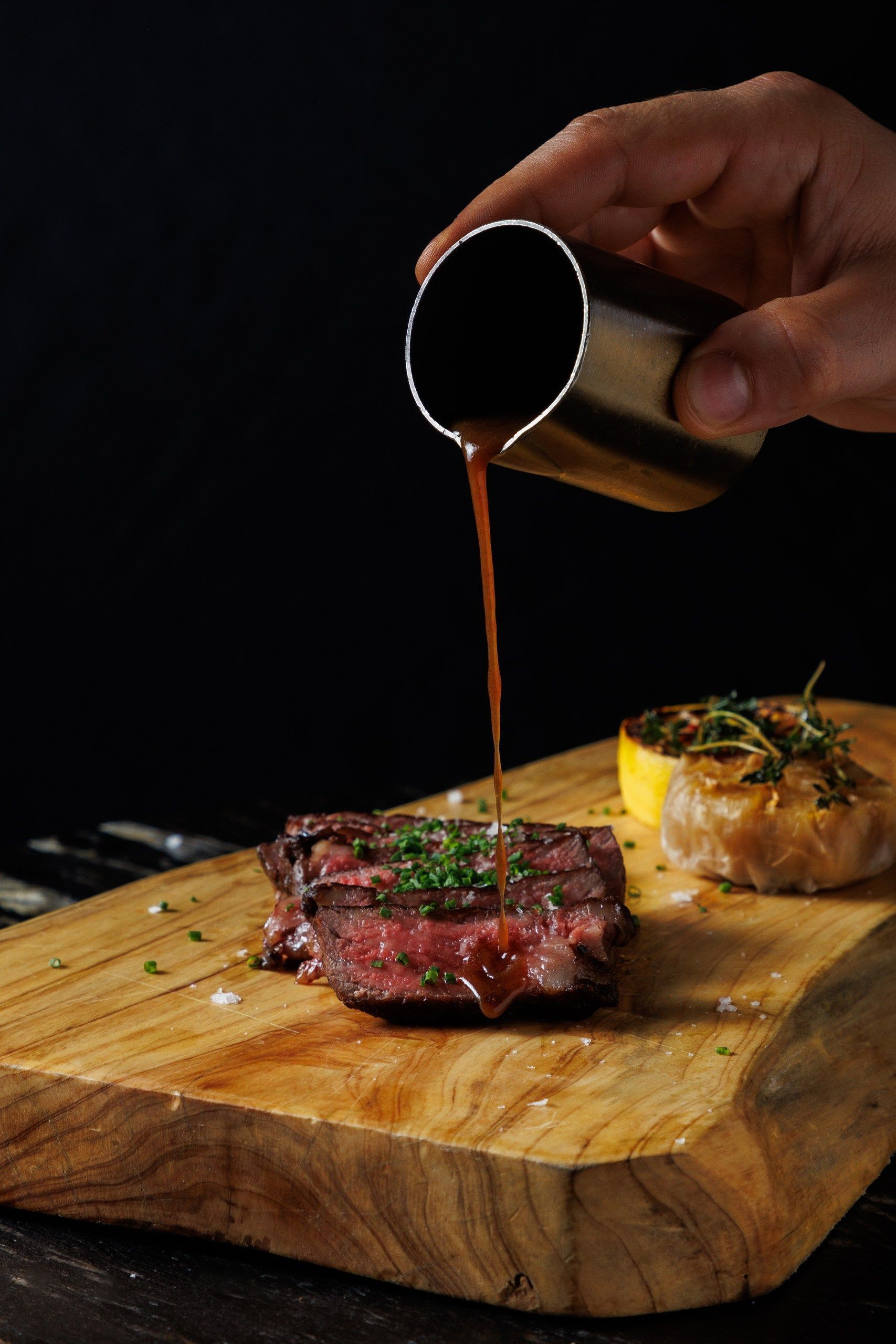 Sauce being poured over sliced steak garnished with herbs on a wooden board, accompanied by roasted vegetables.