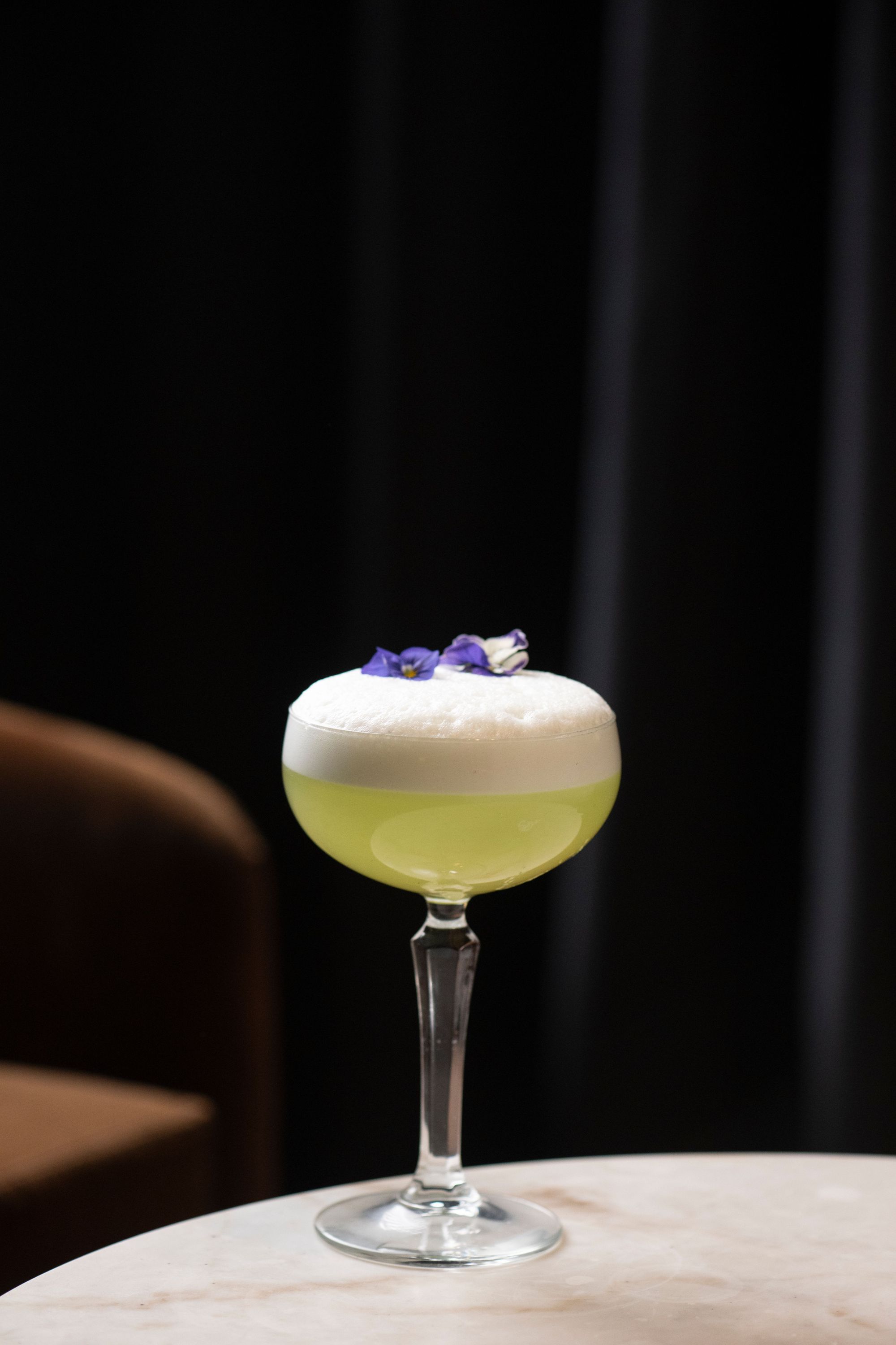 Frosty cocktail with foam and purple flower garnish served in a coupe glass on a marble table.