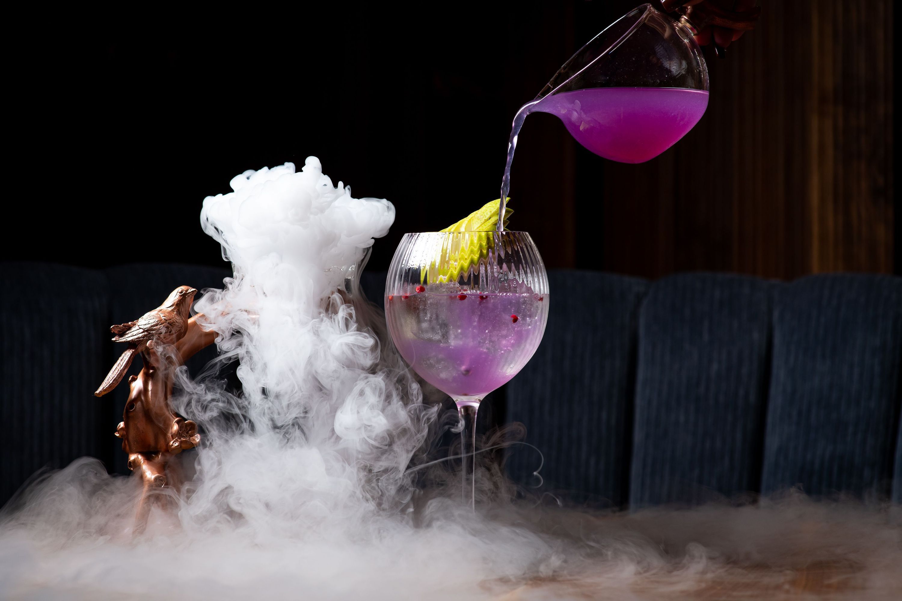 Purple cocktail being poured into a glass with lemon garnish, surrounded by decorative mist and a bird sculpture nearby